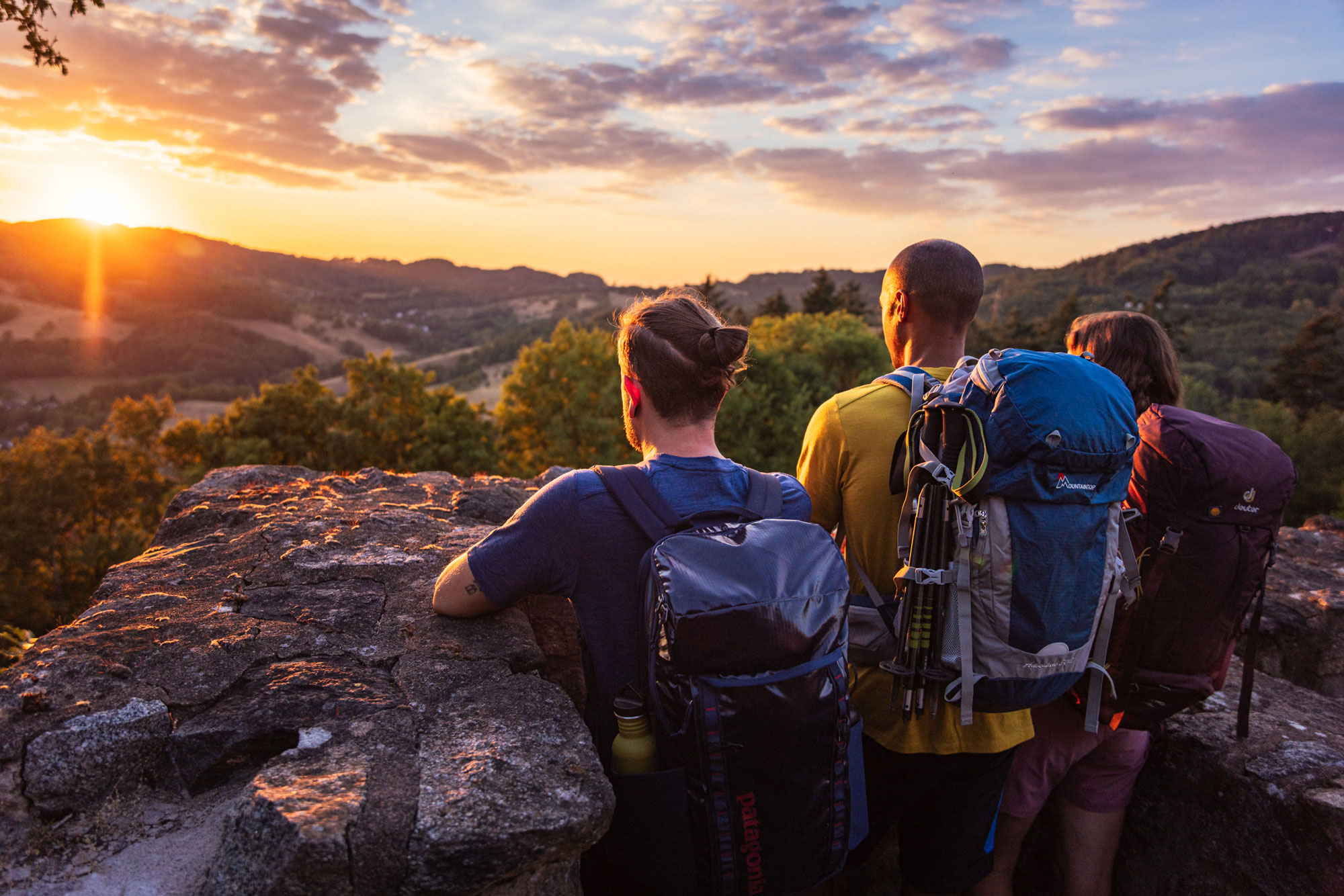 3 Wandernde stehen auf einem Felsen und blicken in den Sonnenuntergang